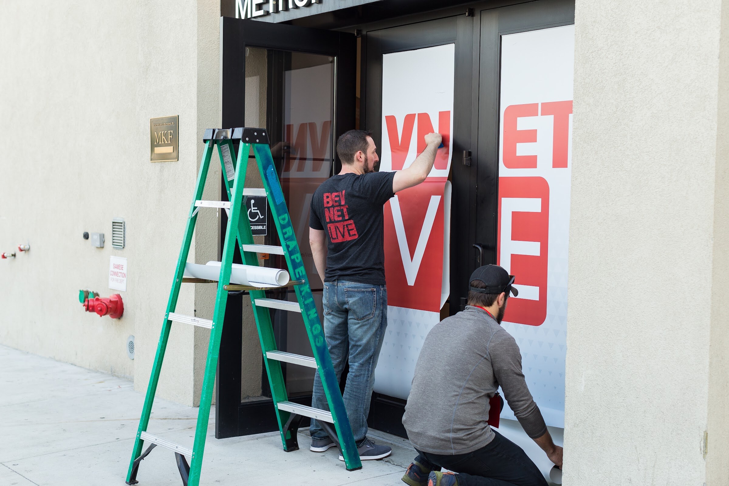 Two men applying red and white BEVNET LIVE vinyl to glass doors, one standing in a BevNET Live T-shirt beside a green ladder and one kneeling.