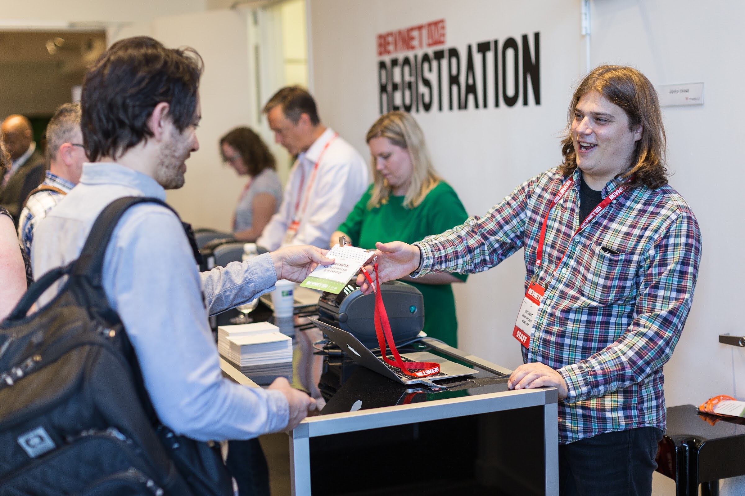 Smiling man in a plaid shirt and red STAFF lanyard handing a badge to an attendee with a backpack, in front of a BevNET Live Registration sign.