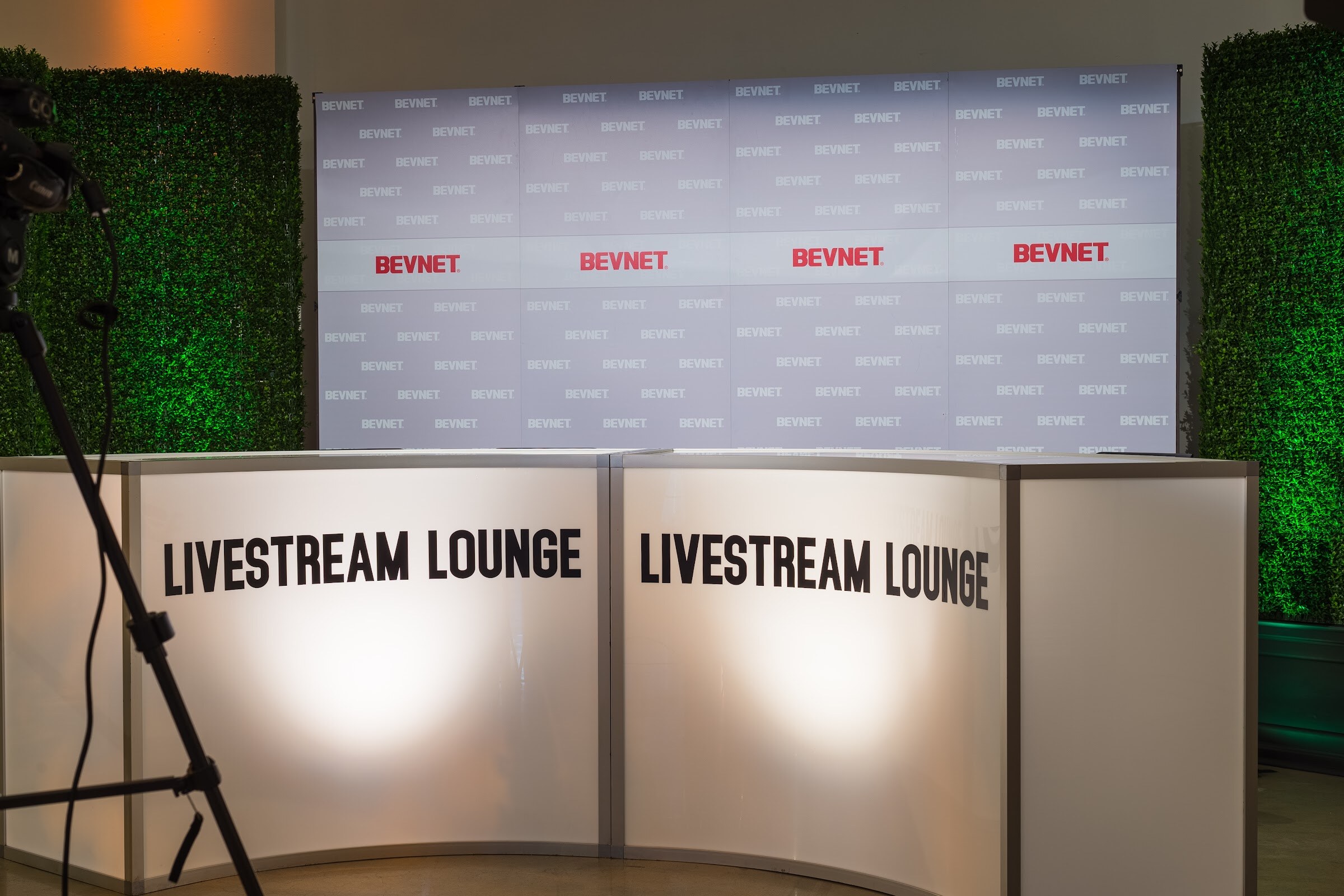 Empty white curved desk reading LIVESTREAM LOUNGE in front of a BEVNET logo wall and green-lit hedge walls, with a camera tripod at left.