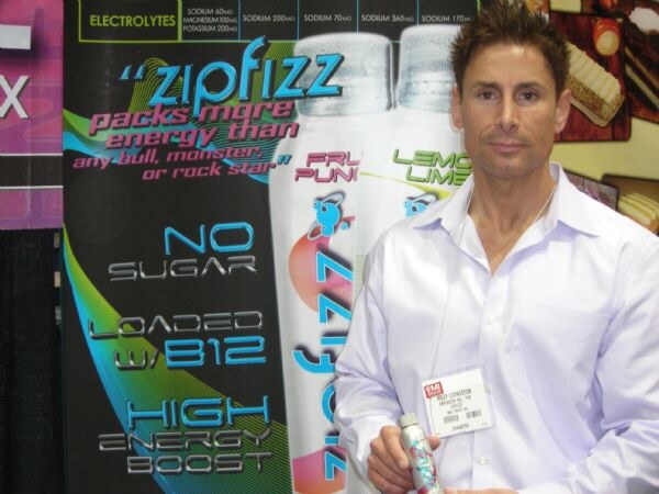 A man in a light button-up shirt with a name badge holds a Zipfizz bottle in front of a Zipfizz banner listing no sugar, B12 and high energy boost.
