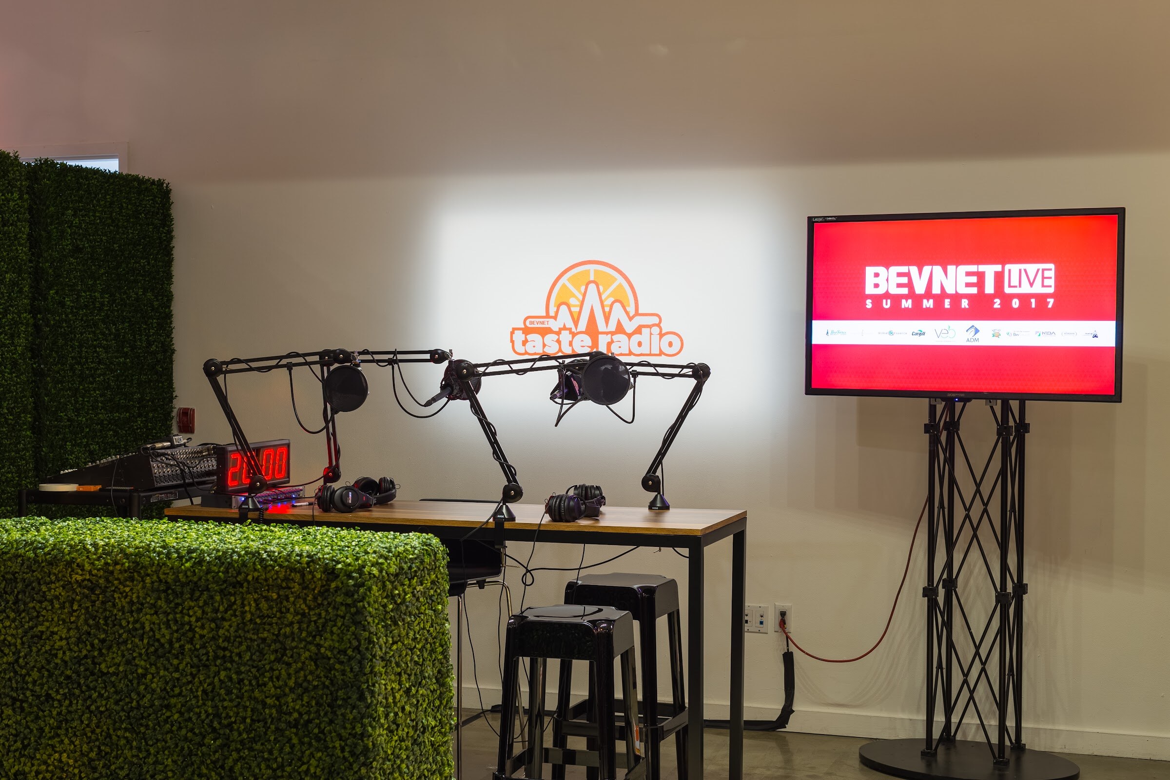 Empty wooden table with boom-arm microphones and headphones in front of a Taste Radio logo on a wall, beside a screen reading BEVNET LIVE SUMMER 2017.