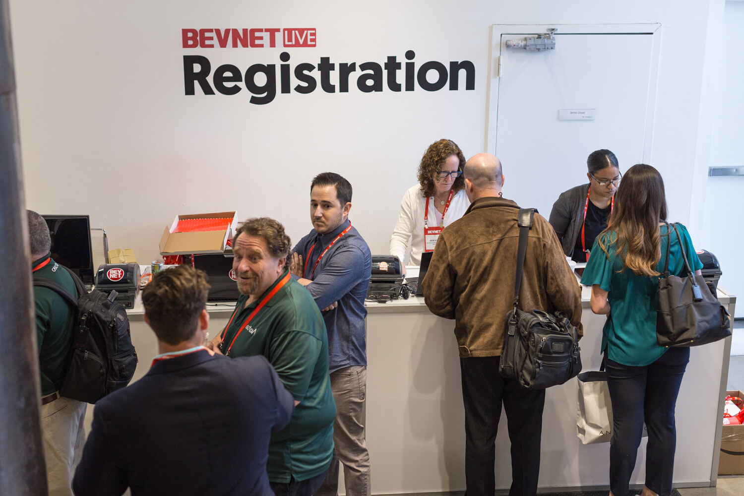 People wearing lanyards stand at a counter below a BevNET Live Registration sign while staff check them in.