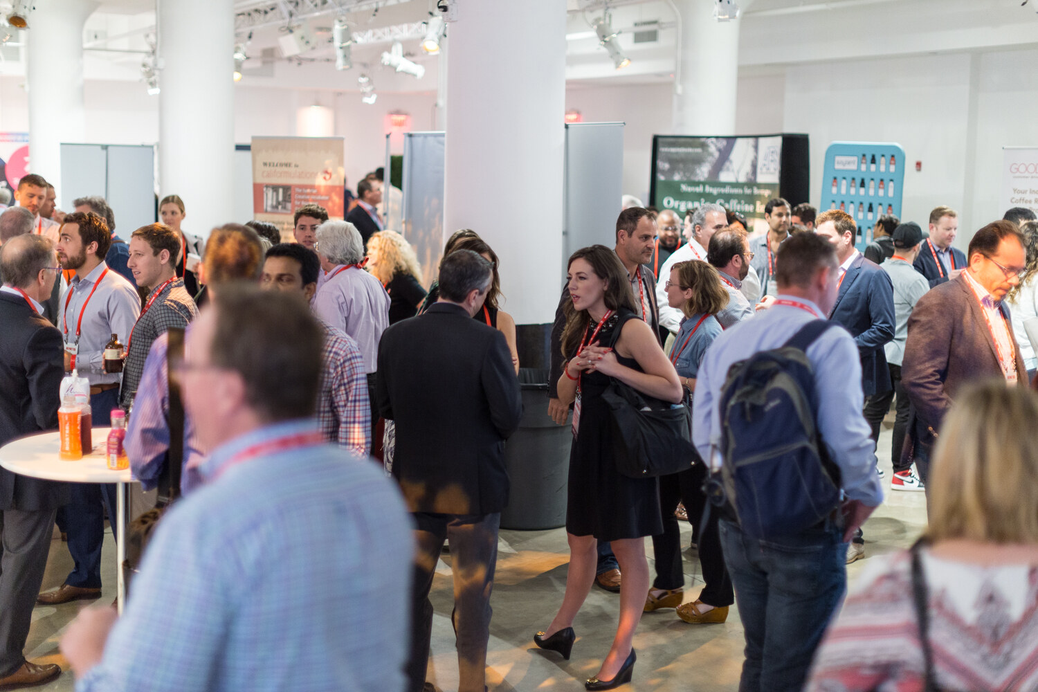 A crowded room of attendees in lanyards standing and talking among white columns and exhibitor banners.