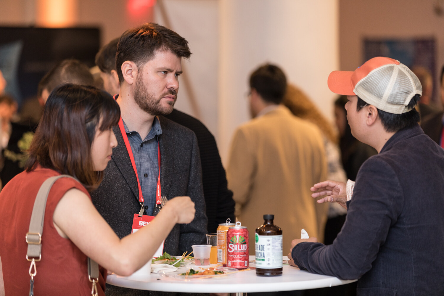 A bearded man and a woman talk with a man in a trucker cap at a white cocktail table with Salud canned drinks on it.