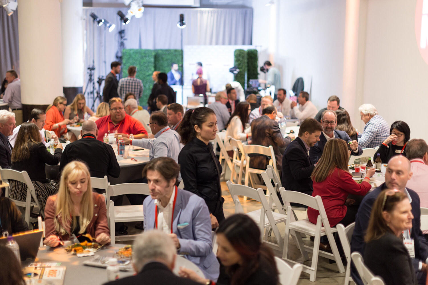 Wide view of attendees seated at round tables with white chairs in a large room with columns and a studio set in the back.
