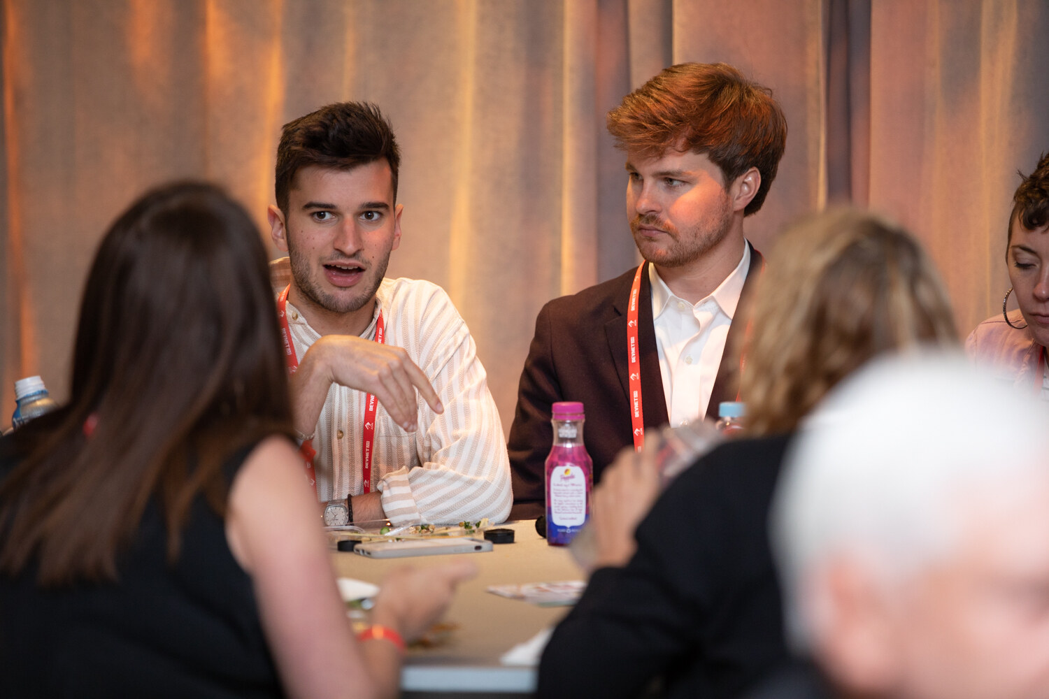 Two young men in lanyards talk at a table, with a woman and a white-haired man in the foreground.
