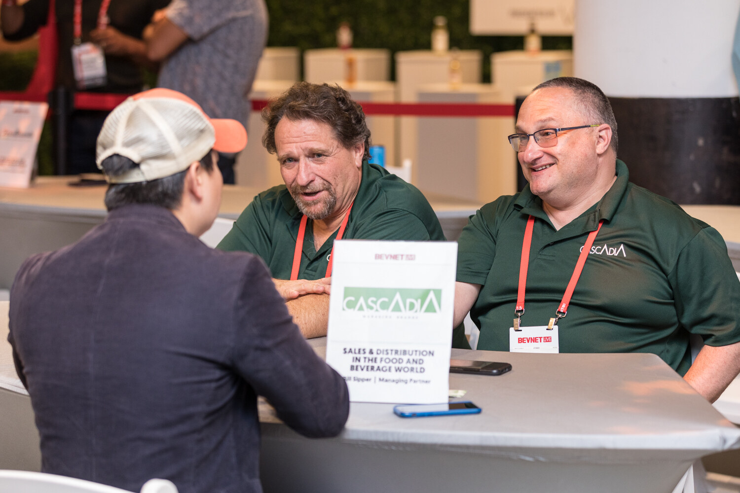 Two men in green polos and lanyards talk with a man in a cap at a table with a Cascadia sales and distribution sign.