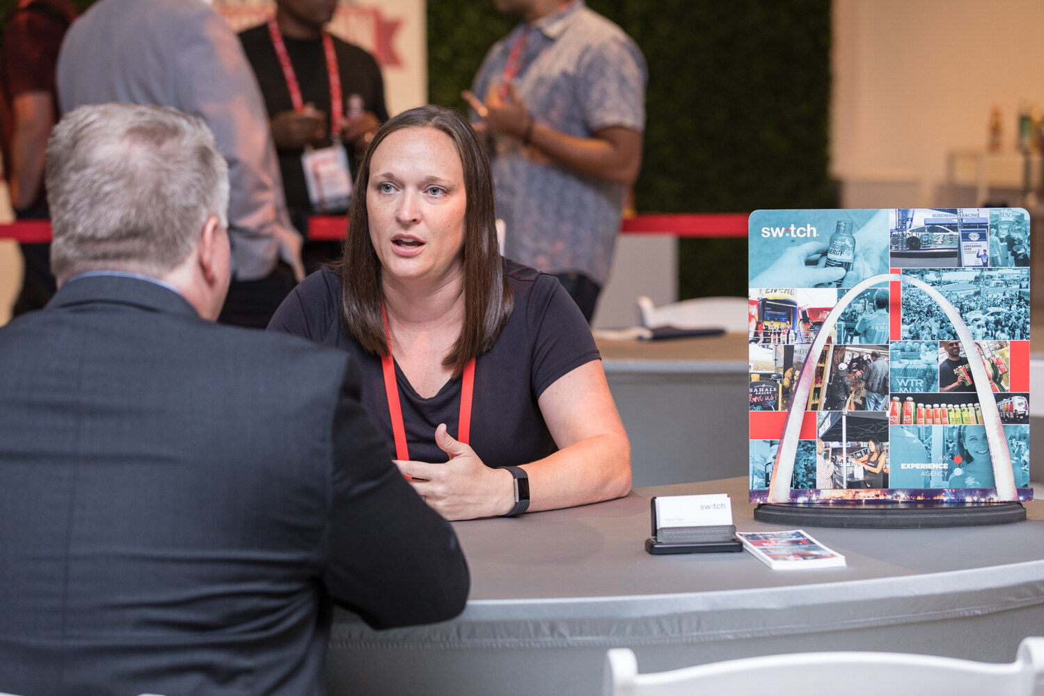 A woman in a black top talks with a man in a gray suit across a table with a business card holder and a promotional display.