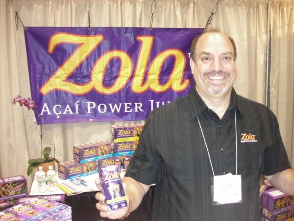 A smiling man in a black shirt with a Zola logo holds a Zola Açaí Power Juice box in front of a purple Zola banner and stacked product boxes.
