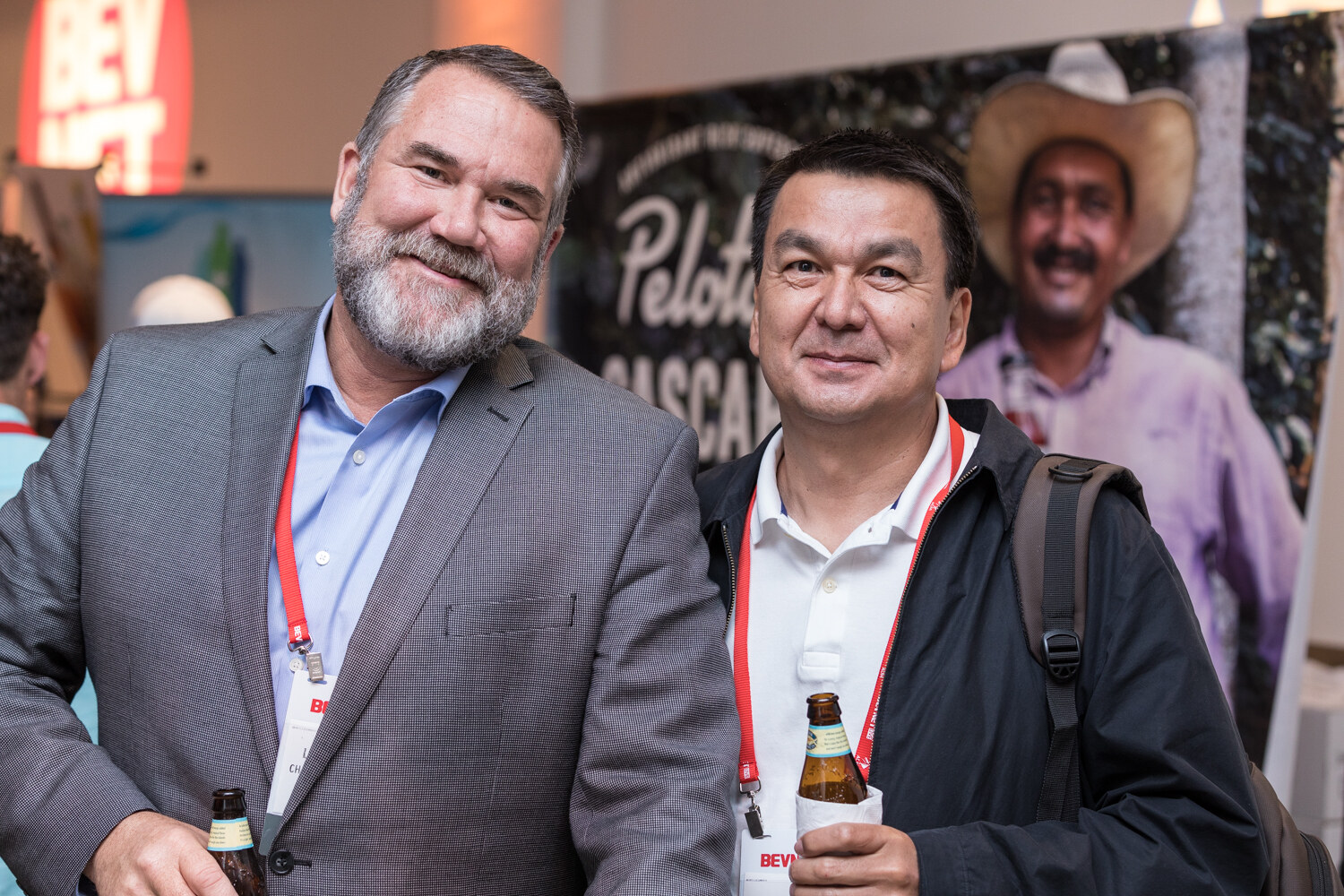 Two men with lanyards and bottles of beer smile at the camera in front of a banner with a Peloton Cascara Tea image.