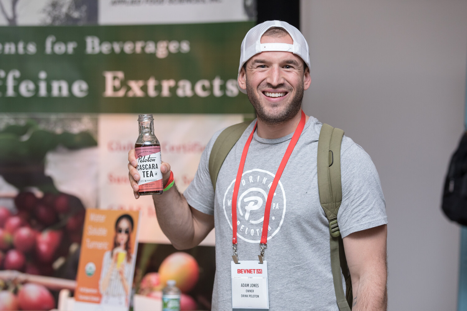 A smiling man in a backwards cap and Peloton t-shirt holds a bottle of Peloton Cascara Tea in front of an Applied Food Sciences banner.