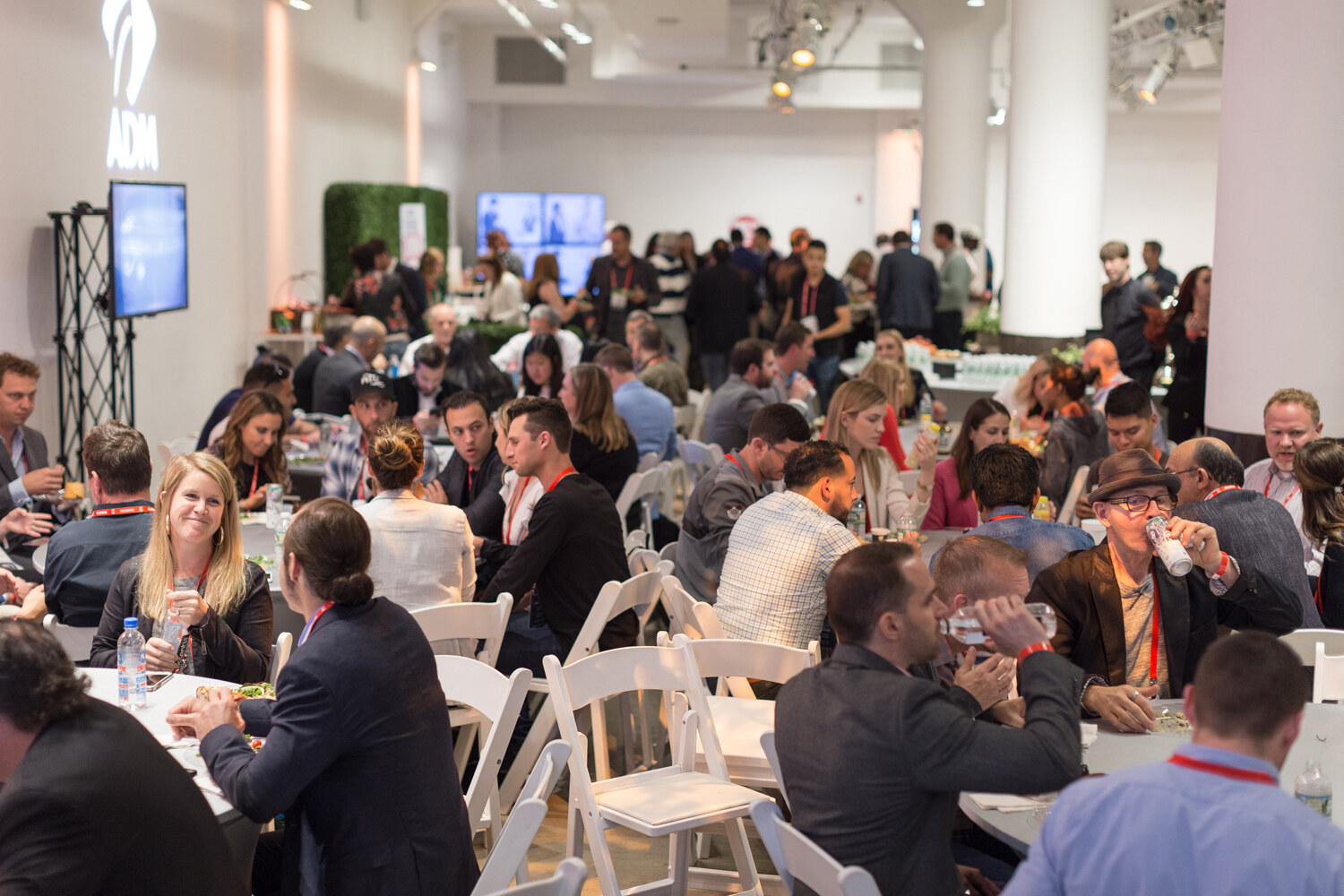 Wide view of many seated attendees at round tables in a large room with columns and an ADM sign.