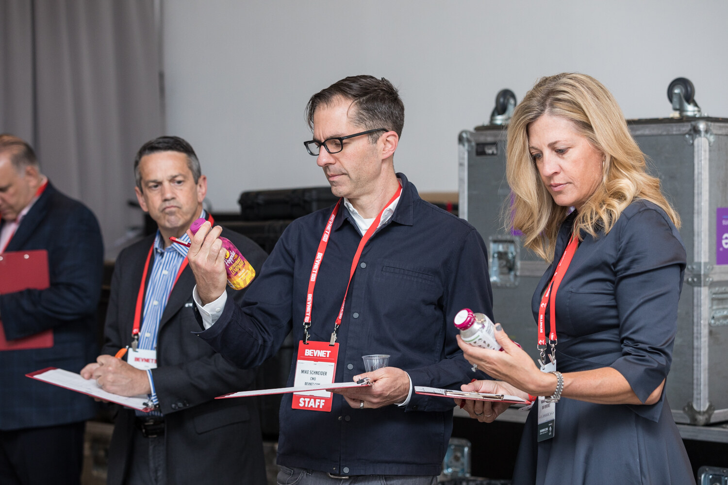 Three people wearing lanyards hold bottles and clipboards backstage while a fourth judge looks on.