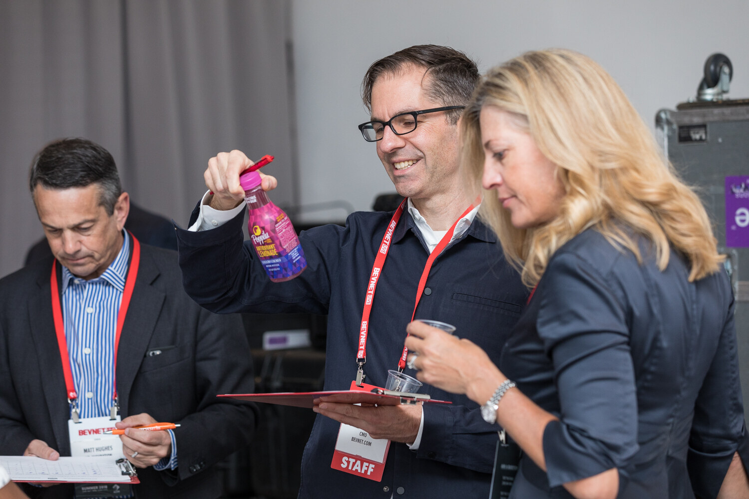 A man in glasses holds a pink bottle with a purple cap while a blonde woman and another man with clipboards look at product.