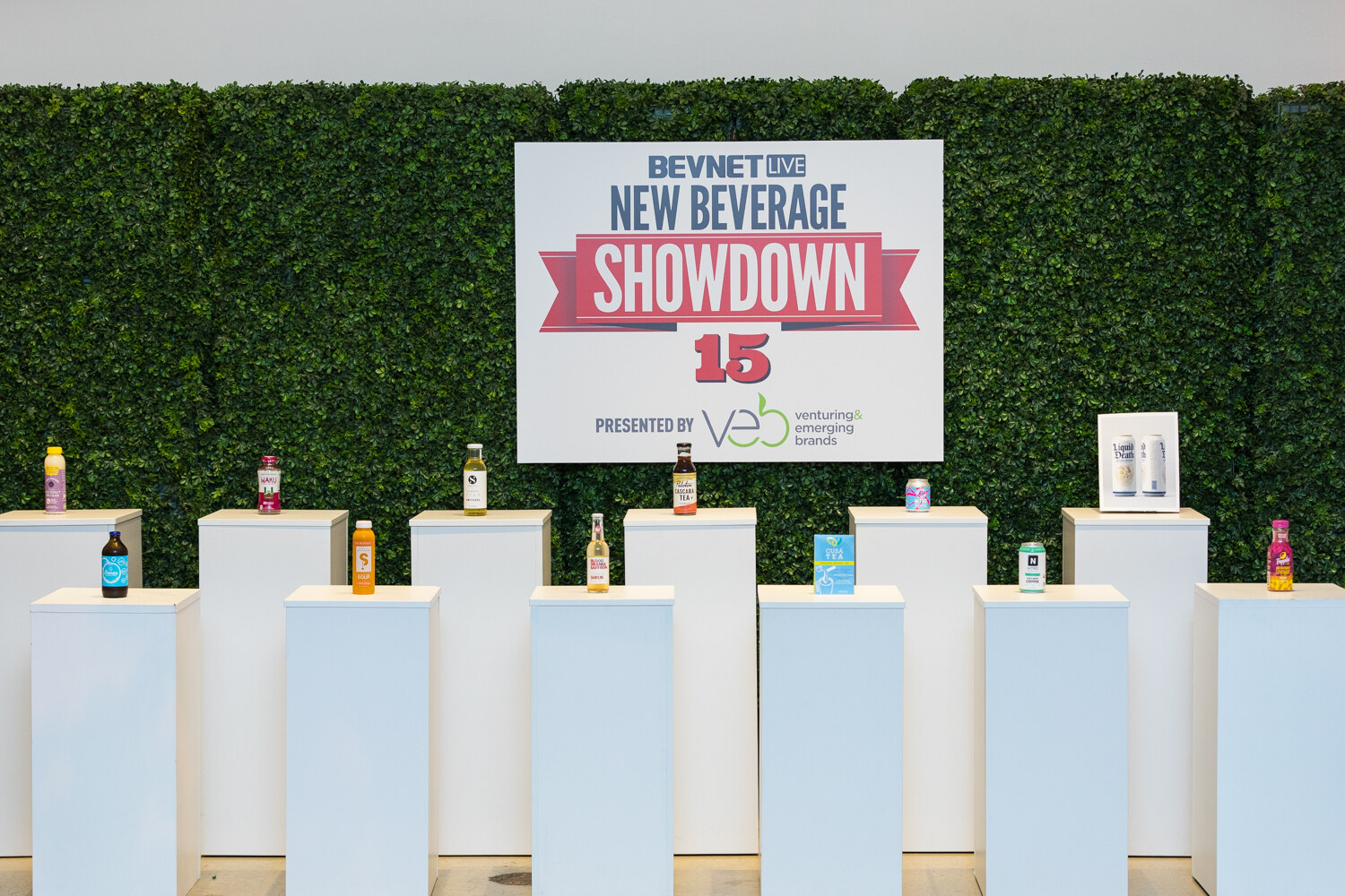 A New Beverage Showdown 15 sign presented by Veb on a hedge wall above rows of white pedestals, each holding a bottle or can.
