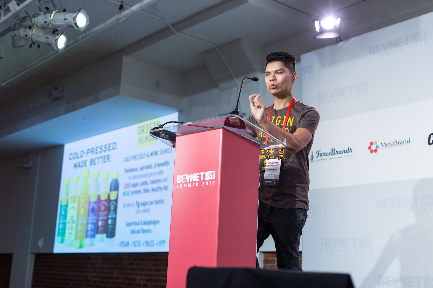 A man in a brown t-shirt with a lanyard speaks at a red lectern beside a screen reading Cold-Pressed Made Better.