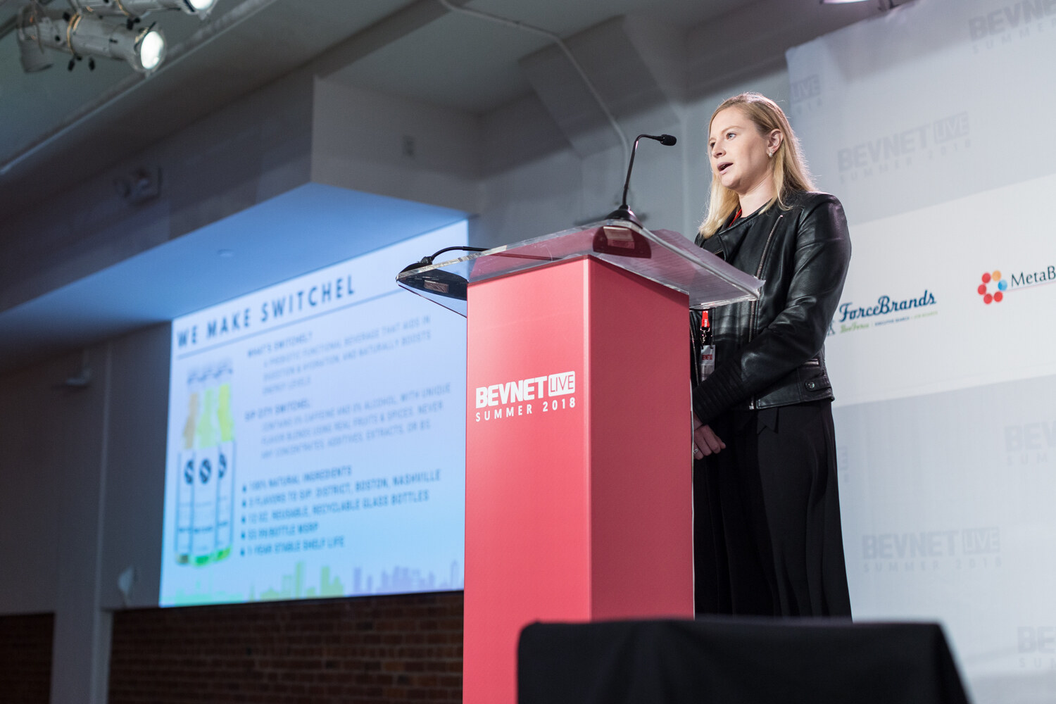 A woman in a black leather jacket speaks at a red lectern next to a projected slide titled We Make Switchel.