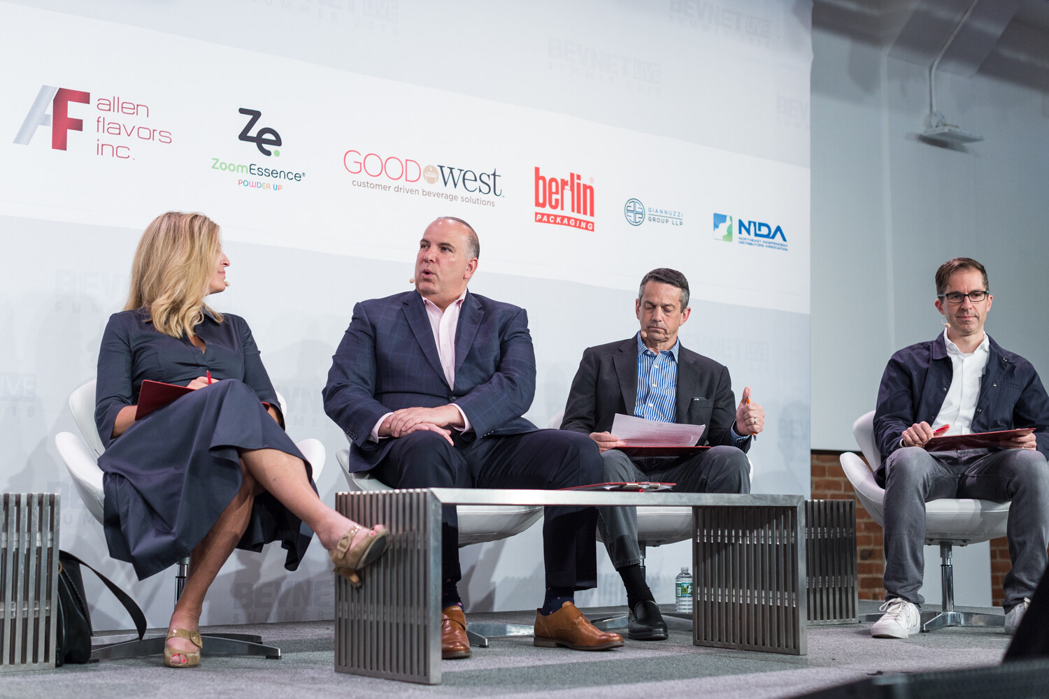 Four people seated on stage in white chairs holding folders, in front of a sponsor backdrop with Allen Flavors, ZoomEssence and Good West logos.