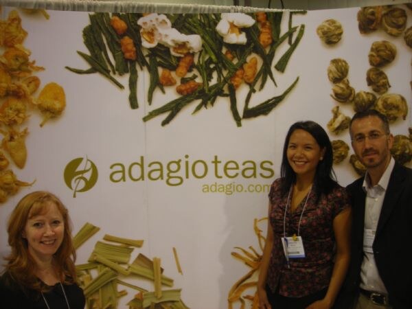 Two women and a man smile in front of an adagioteas.com backdrop decorated with dried tea leaves, flowers and tea balls.