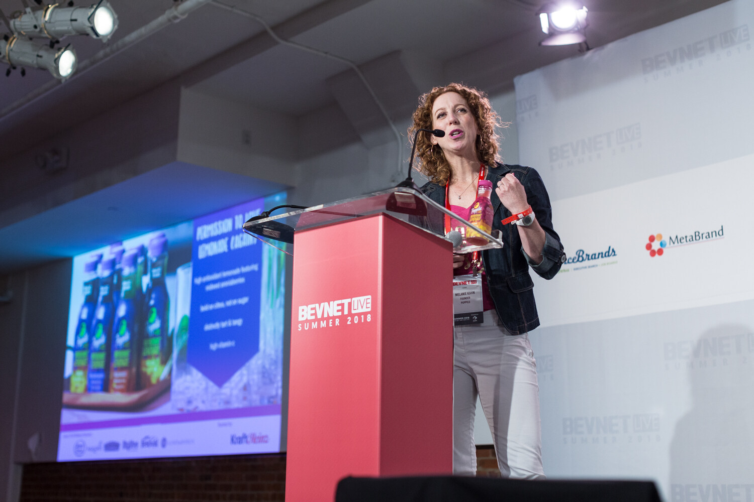 A woman with curly hair speaks at a red lectern with a pink bottle in hand, beside a large screen showing bottled drinks.