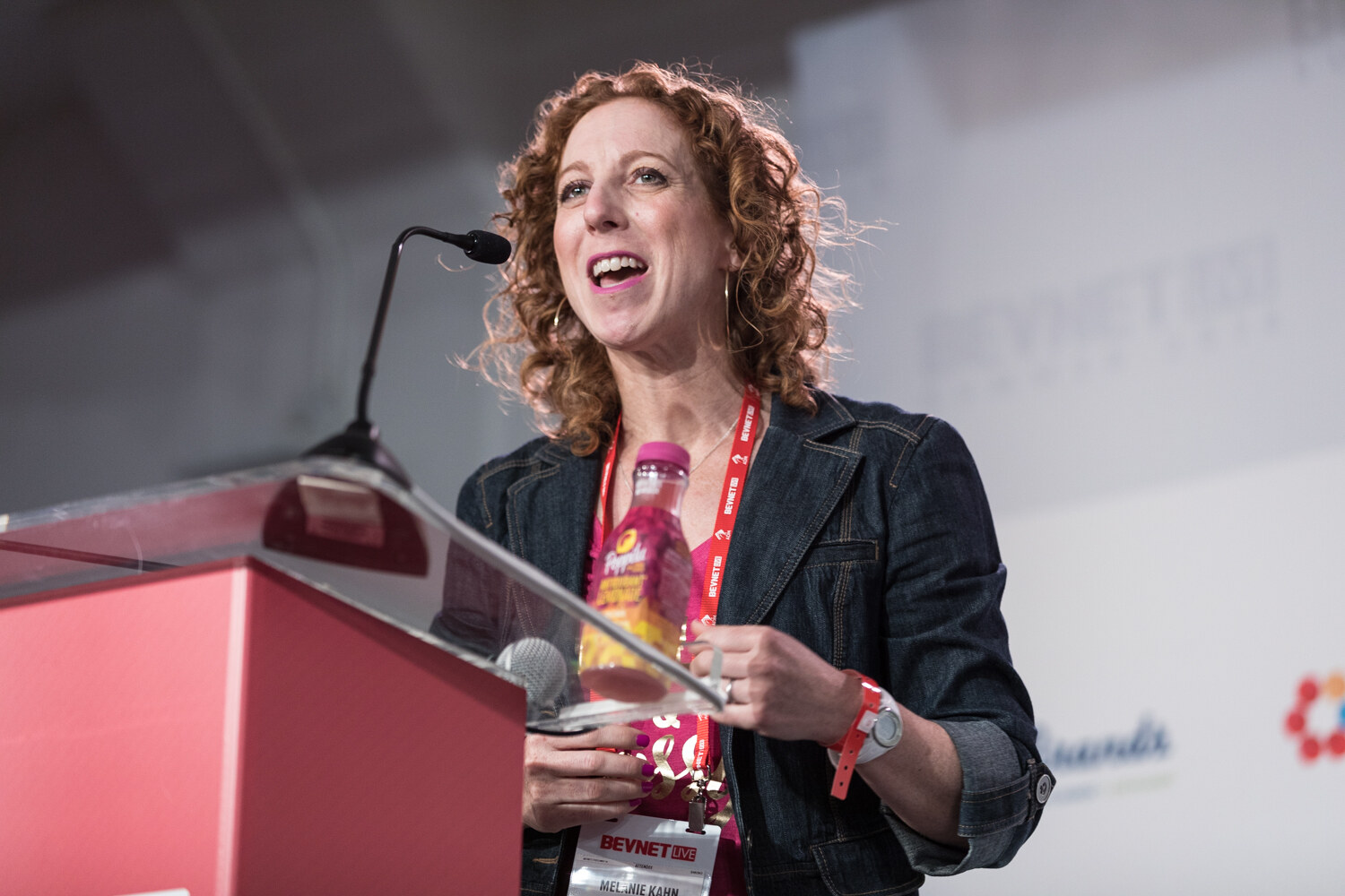 A woman with curly hair smiles while speaking at a lectern, holding a pink and yellow bottle in her hands.
