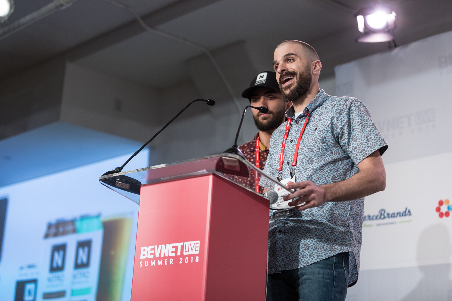 A bearded man in a patterned shirt speaks at a red lectern while a second man in a black Nitro cap stands behind him.
