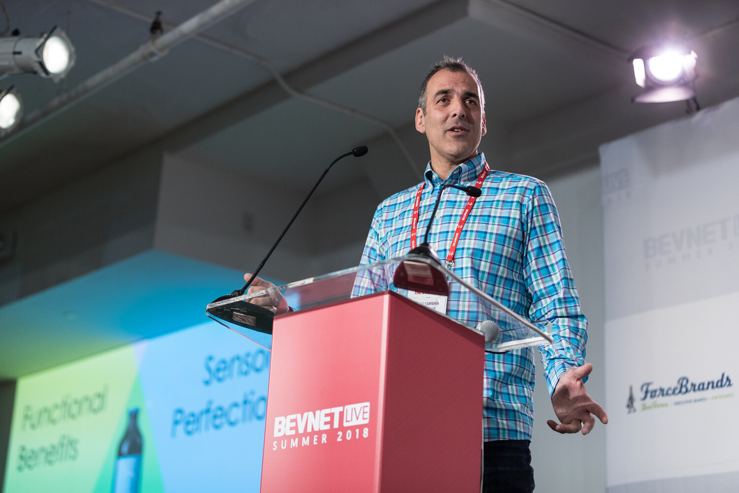 A man in a blue plaid shirt gestures at a red lectern beside a slide mentioning functional benefits.