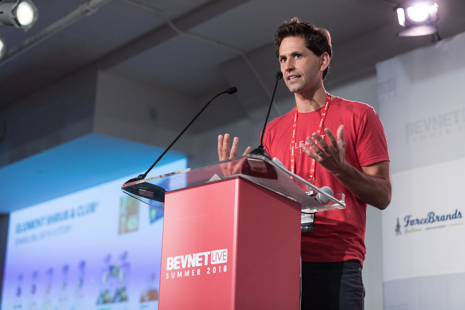 A man in a red t-shirt with a lanyard gestures with both hands at a red lectern beside a slide reading Element Shrub & Club.