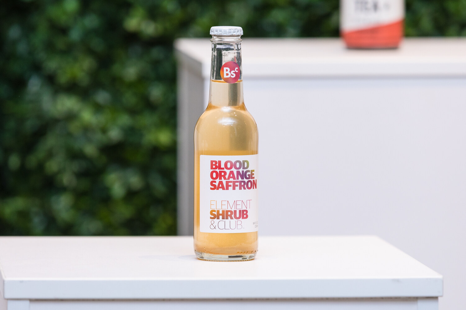 A clear glass bottle labeled Blood Orange Saffron, Element Shrub & Club, on a white pedestal in front of a green hedge wall.