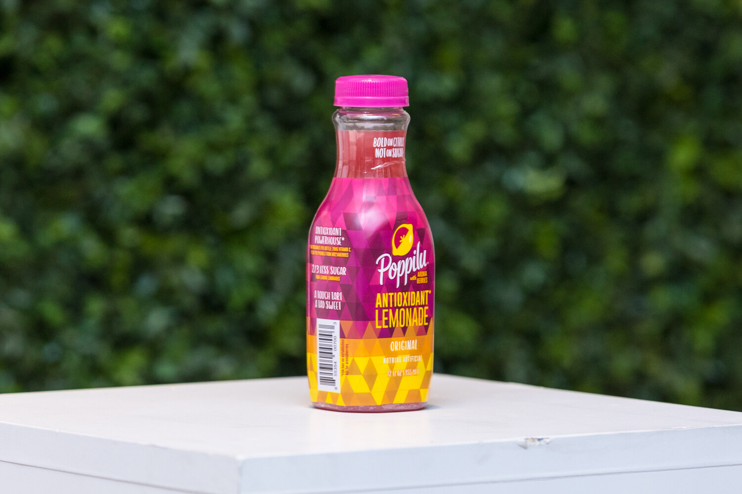 A pink and yellow bottle labeled Antioxidant Lemonade on a white pedestal in front of a green hedge wall.