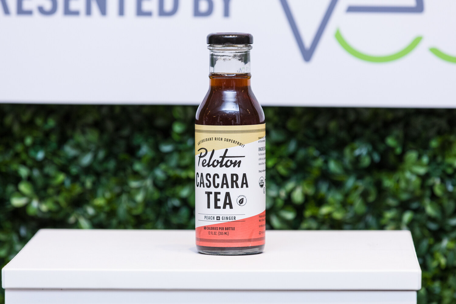 A glass bottle of Peloton Cascara Tea with a red label on a white pedestal under a sign that reads Presented by Veb.