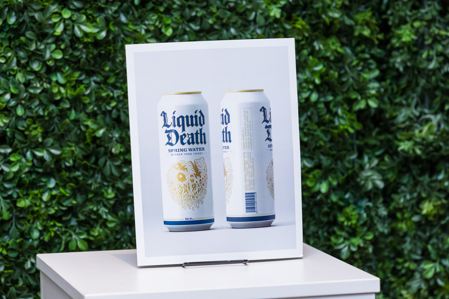 Two white Liquid Death spring water cans in a framed photograph on a white pedestal in front of a hedge wall.