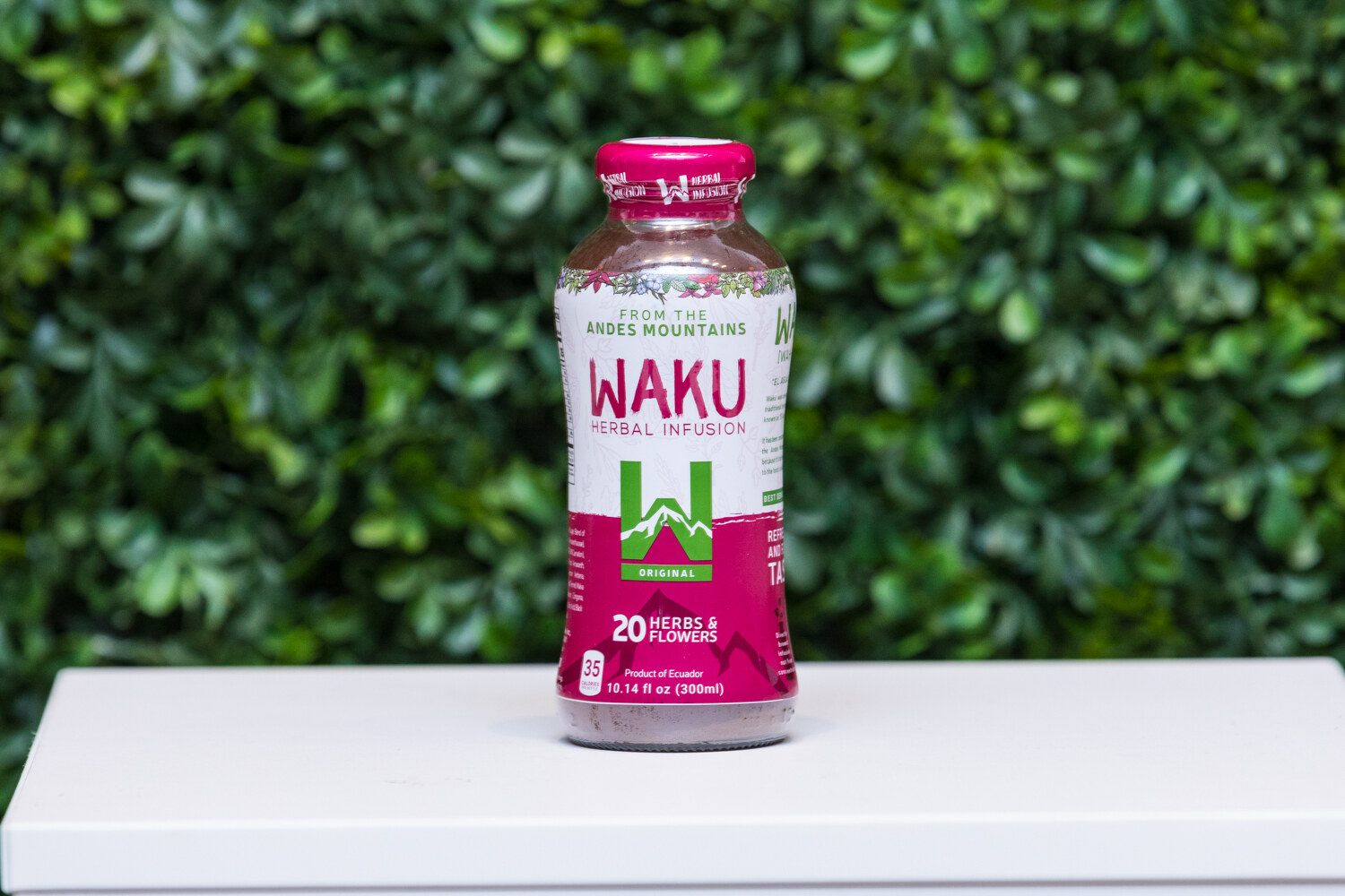 A magenta-labeled bottle of WAKU Herbal Infusion from the Andes Mountains on a white pedestal in front of a hedge wall.