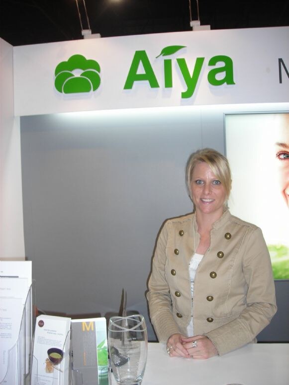 A blonde woman in a tan jacket with gold buttons stands at a white counter beneath a green Aiya sign, with brochures and a glass vase in front of her.