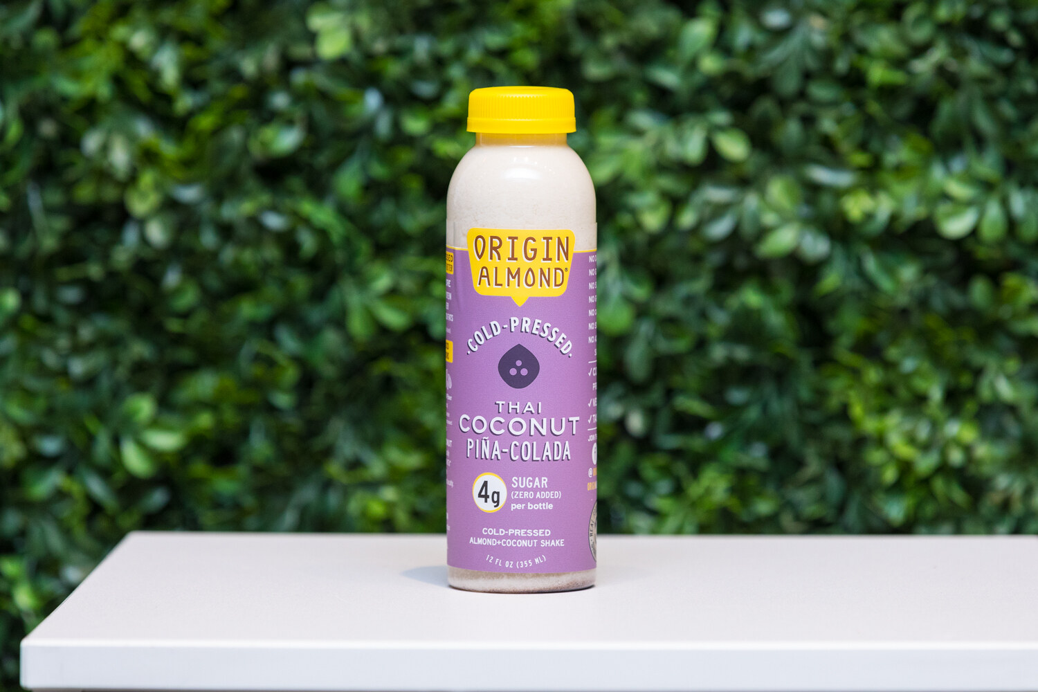 A bottle with a purple label and yellow cap reading Origin Almond, Thai Coconut Pina-Colada, on a white pedestal in front of greenery.