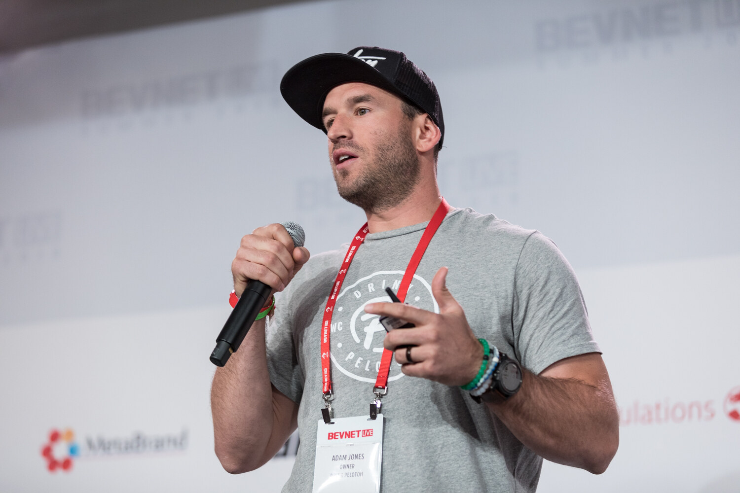 A man in a black cap and gray t-shirt with a lanyard holds a microphone and gestures while speaking in front of the sponsor backdrop.