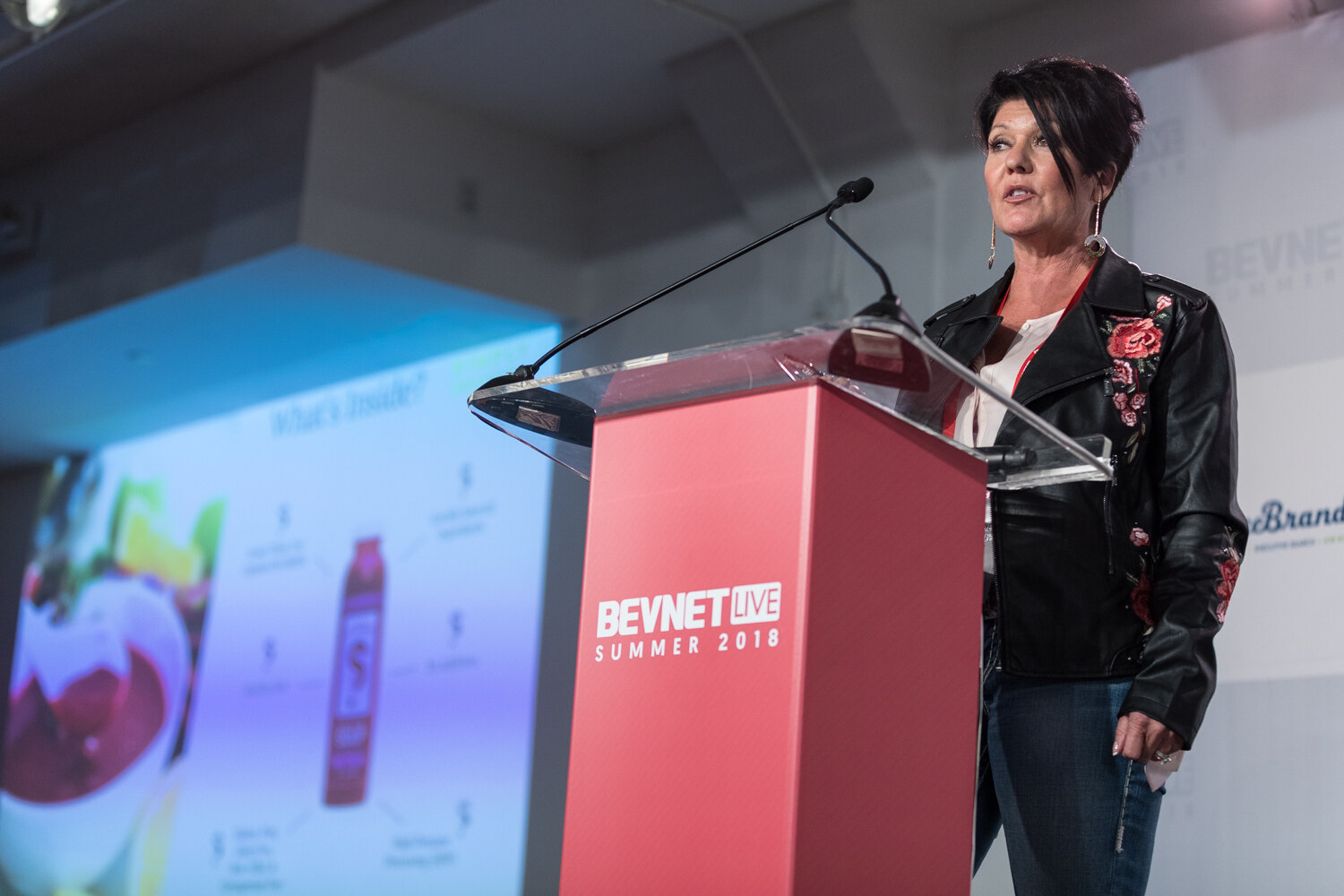 A woman with short dark hair in a black floral-embroidered jacket speaks at a red lectern beside a slide with a bottle graphic.