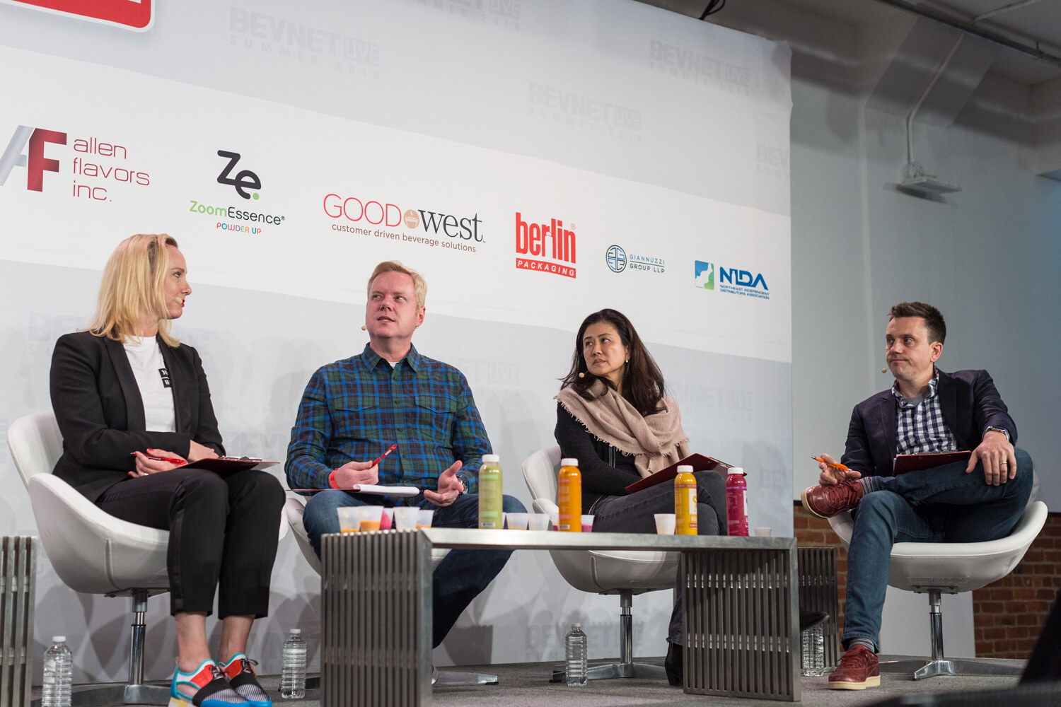 Four judges in white chairs on stage with small sample cups, bottles and clipboards on a metal coffee table.