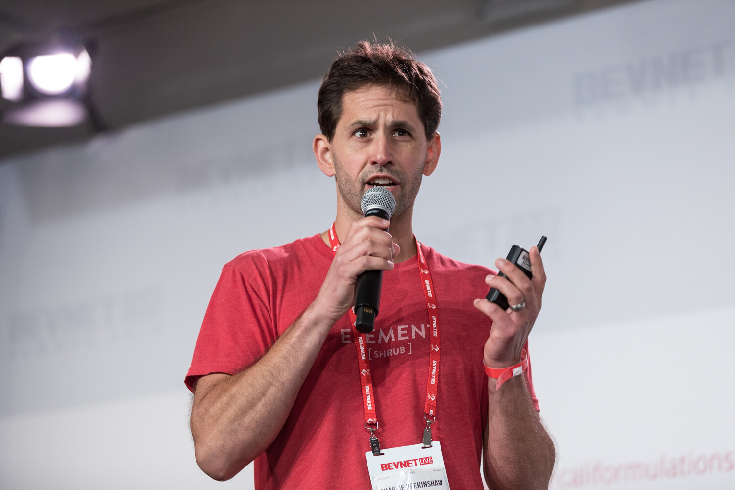 A man in a red Element t-shirt and BevNET lanyard holds a microphone and a clicker while speaking.