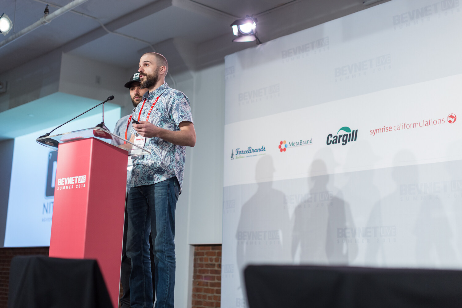 Two men at a red BevNET Live Summer 2018 lectern, one in a patterned shirt and one in a black Nitro cap, with sponsor logos behind them.