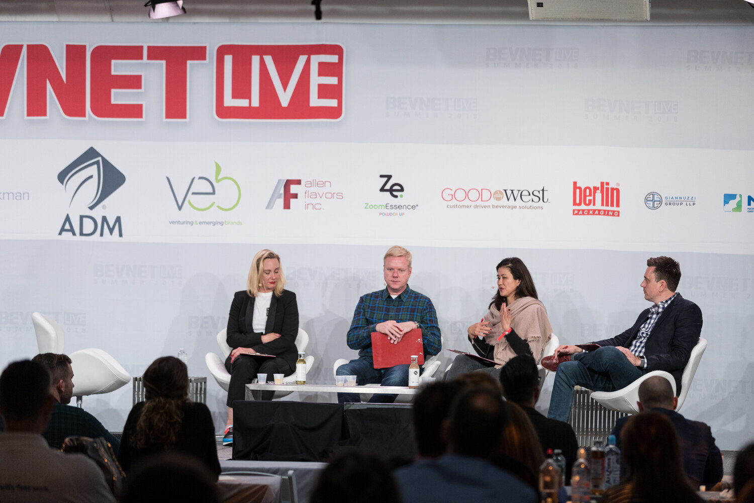 Wide view of four people seated on stage with sample cups on a table in front of the BevNET Live sign and sponsor logos.