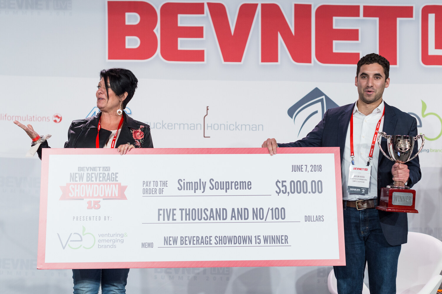 A woman and a man hold a large check reading Simply Soupreme, Five Thousand and No/100, New Beverage Showdown 15 Winner, and a silver trophy.