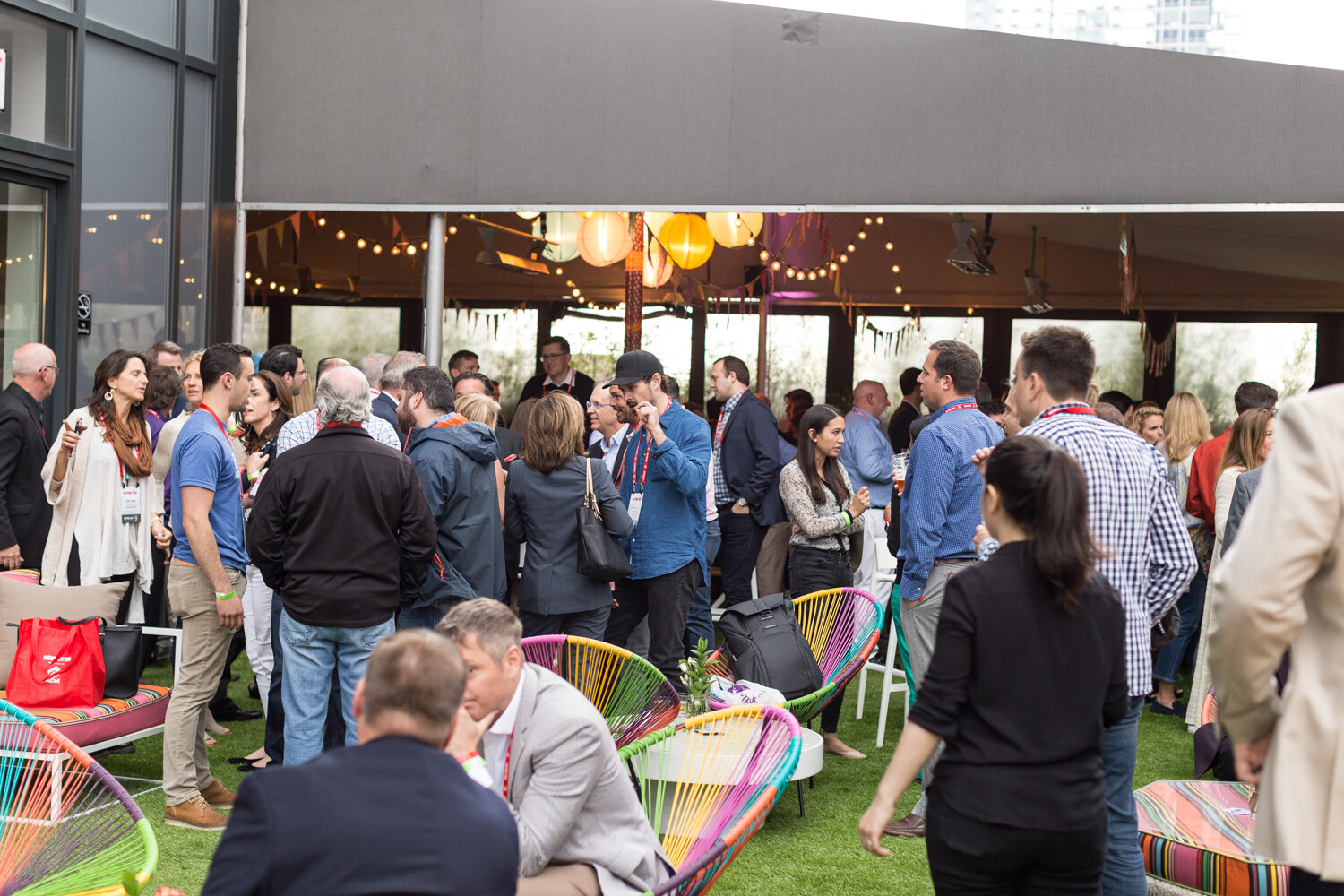 A crowd of people standing and talking on an outdoor terrace with colorful woven chairs and string lights.