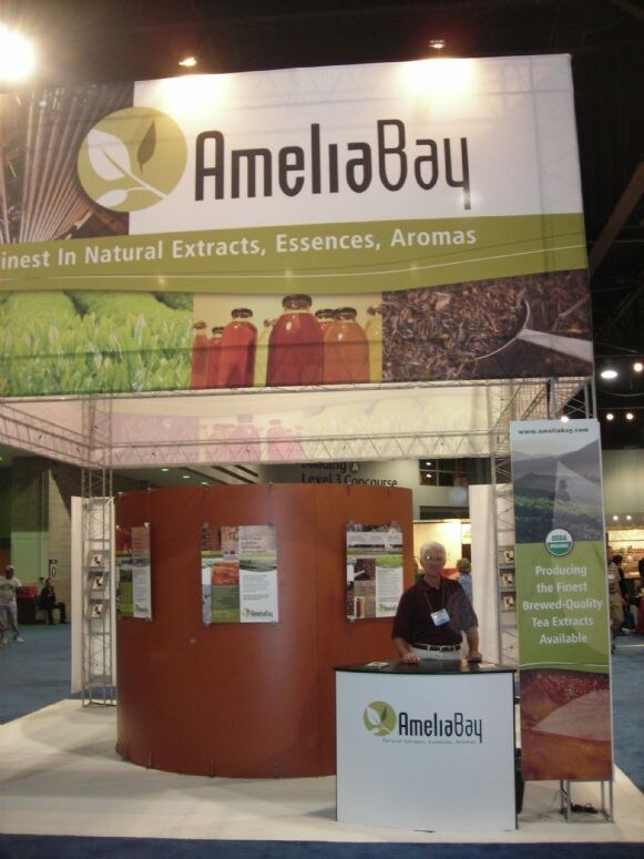 A man stands behind a small counter at the AmeliaBay booth, which has a curved orange display wall with posters and a large banner reading Finest in Natural Extracts, Essences, Aromas.