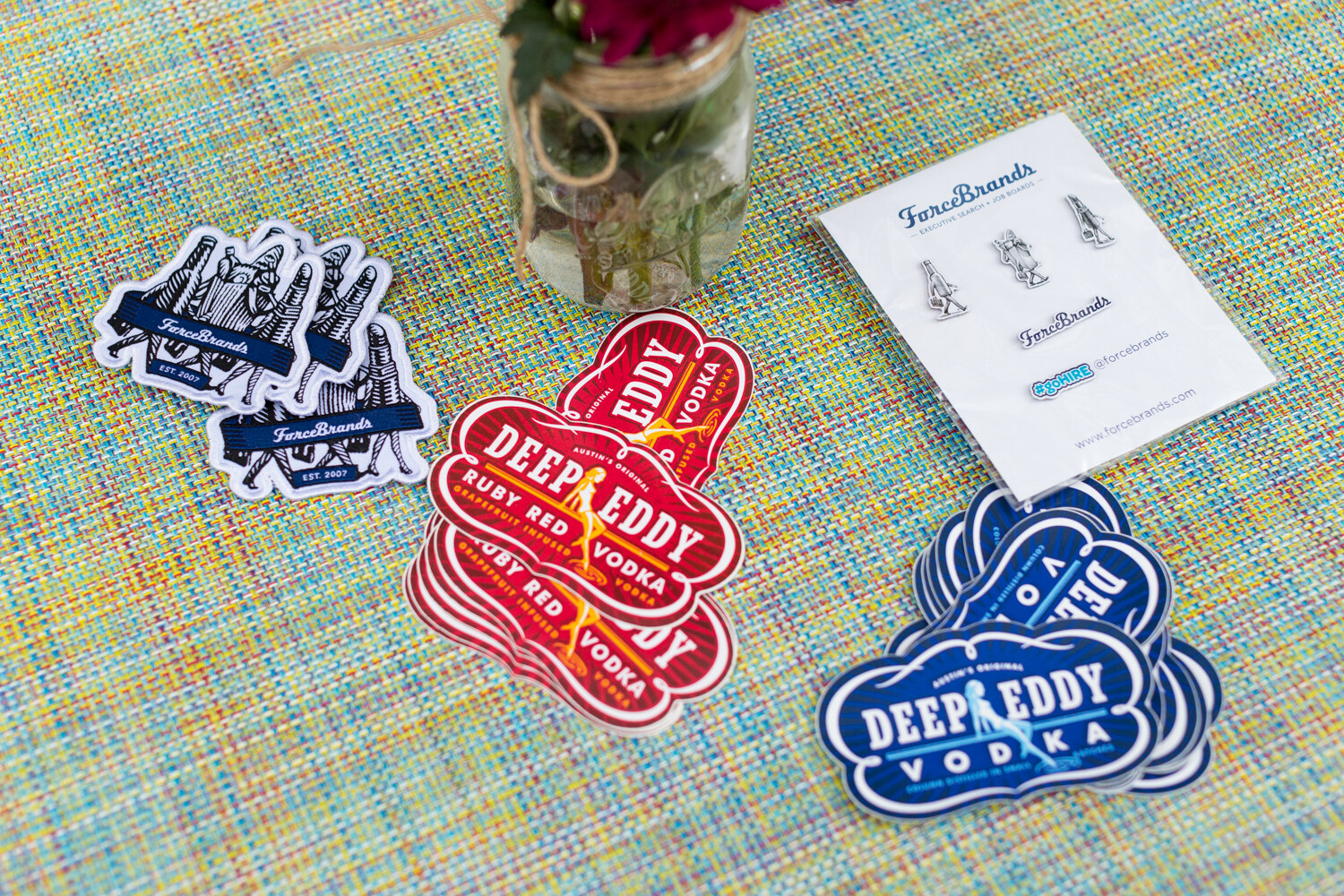 Stickers reading ForceBrands and Deep Eddy Ruby Red Vodka and a ForceBrands pin card on a woven tablecloth beside a small flower jar.