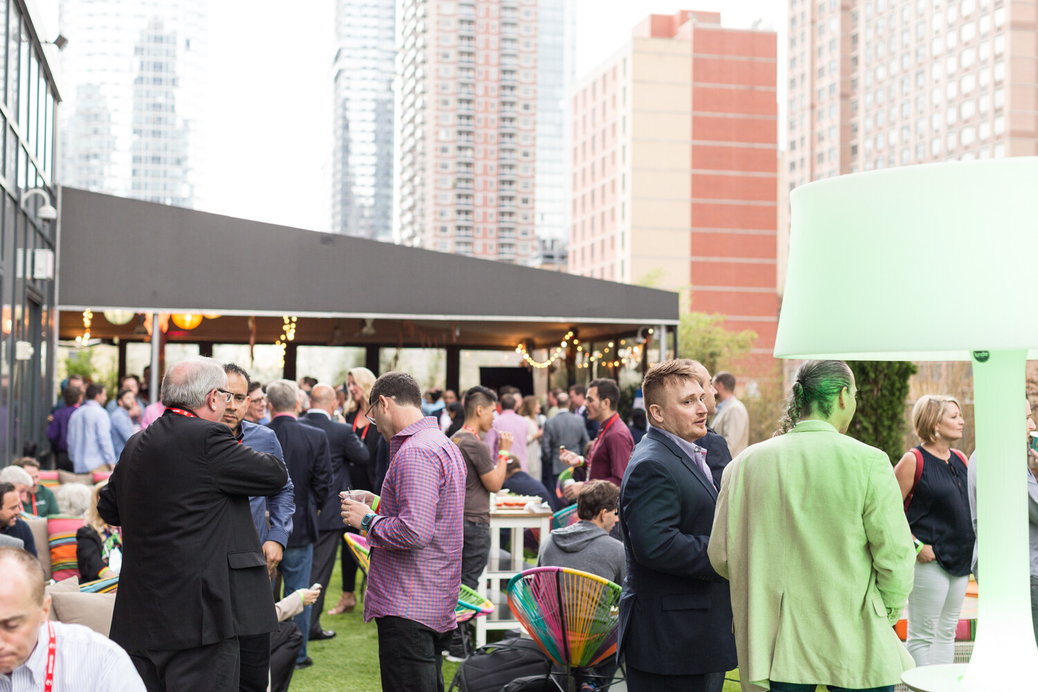 People talk on a terrace with colorful chairs and a green lamp, with apartment towers behind them.