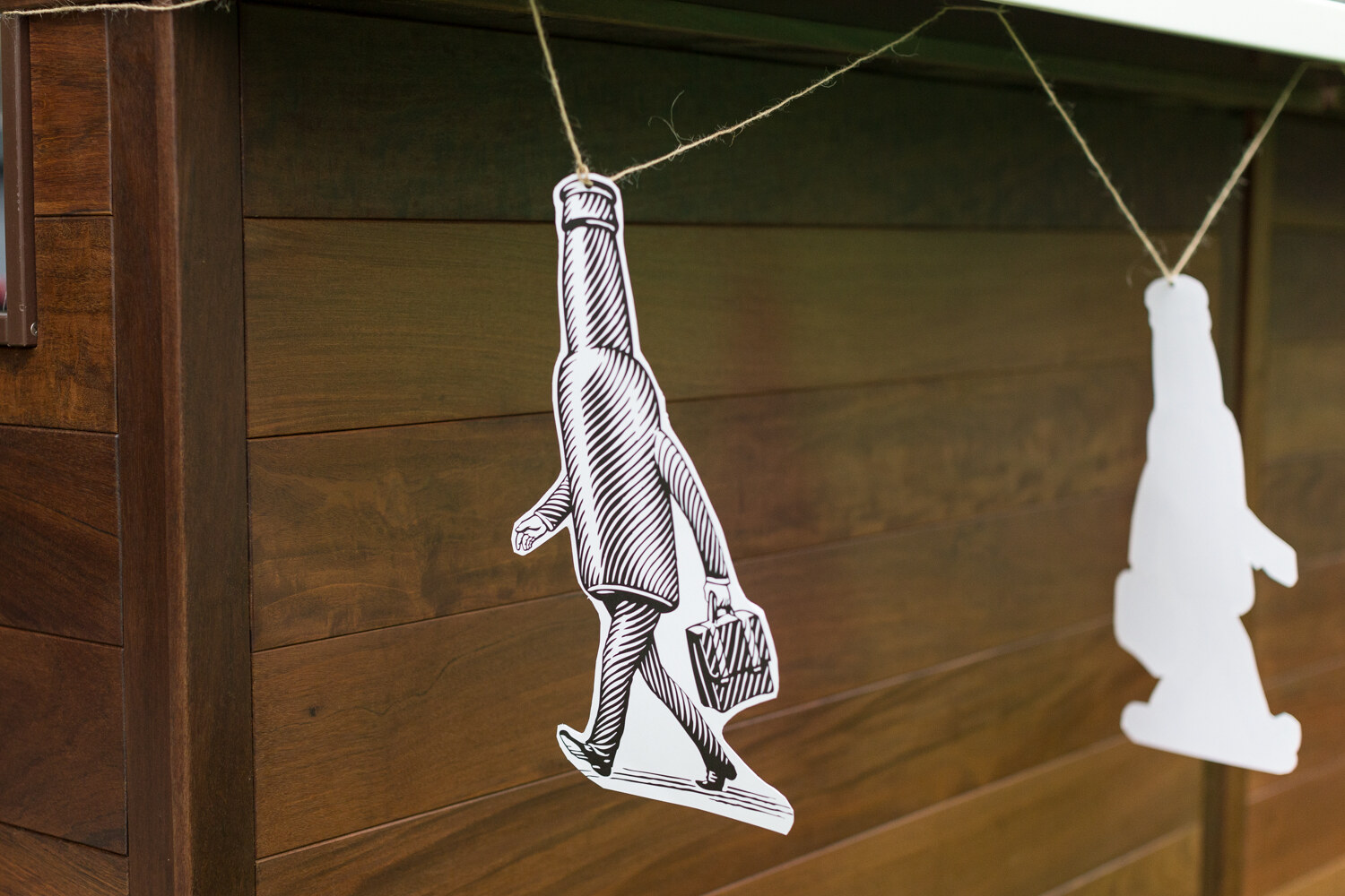 Two paper figures shaped like a walking bottle carrying a briefcase hang from twine on a wood-paneled wall.