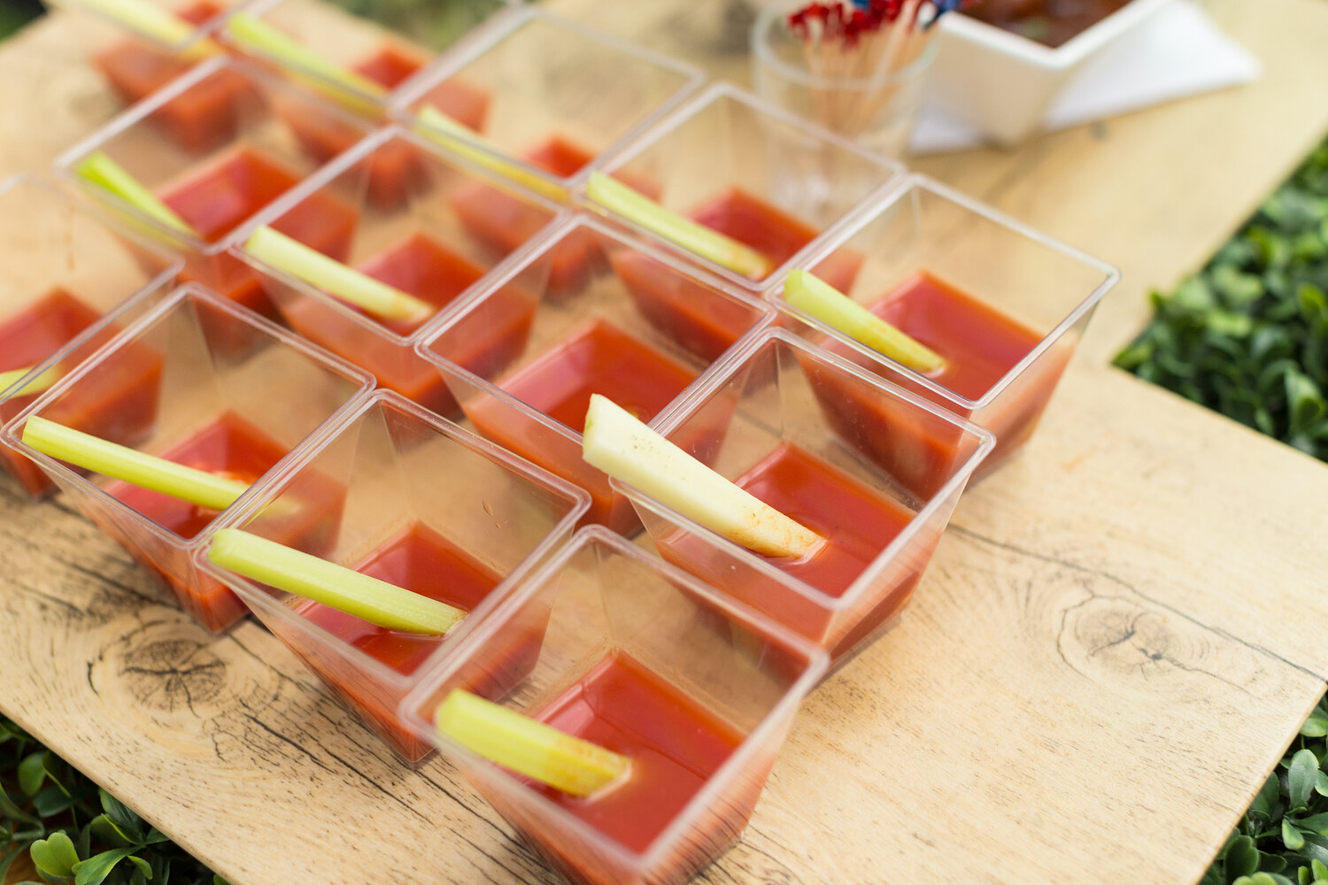 Rows of clear square cups holding a red tomato drink, each with a celery stick, on a wooden board.