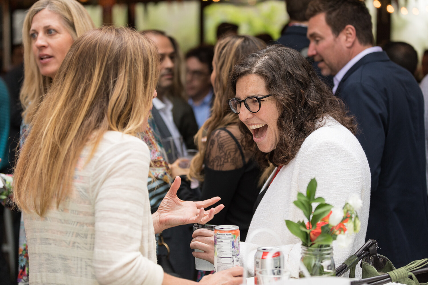 Two women talk and laugh while holding cans, with a vase of flowers and other guests behind them.