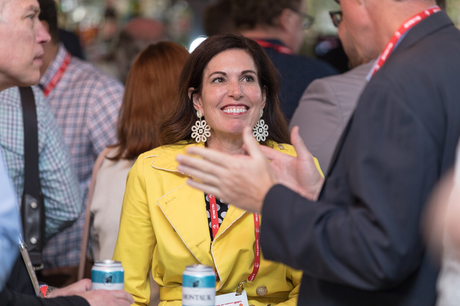 A smiling woman in a yellow jacket and statement earrings with a lanyard, with a man's gesturing hands in the foreground.